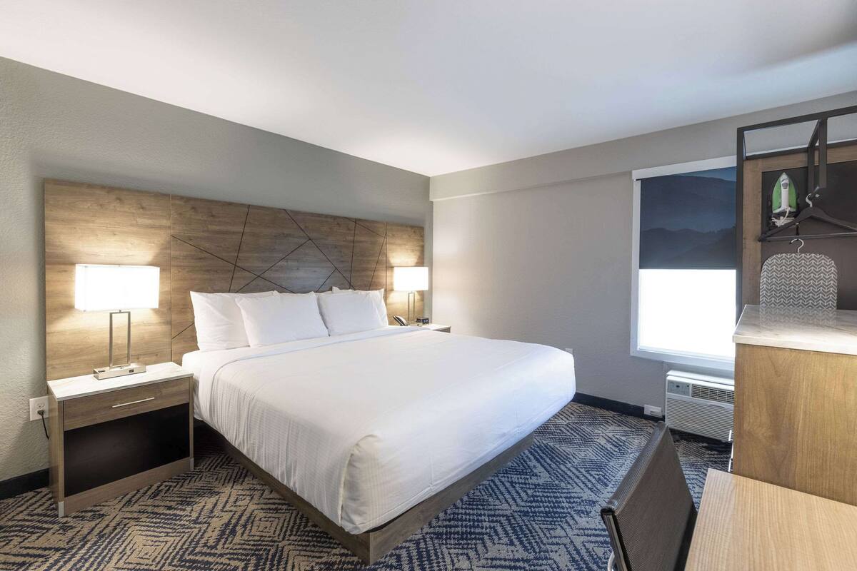 Quarto de hotel com uma cama grande, duas mesas de cabeceira com luminárias, cabeceira de madeira, tapete estampado e uma janela com vista para as montanhas.