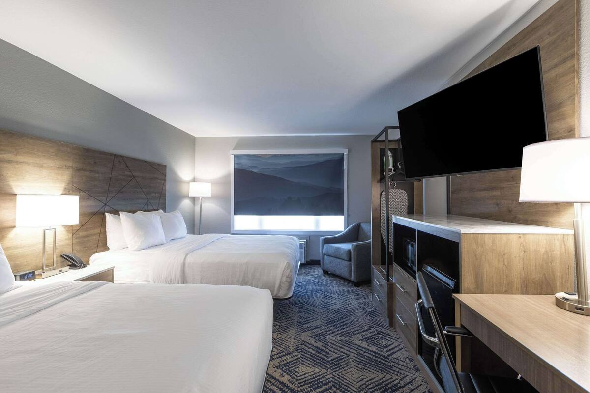 Quarto de hotel com duas camas, uma escrivaninha, uma cadeira, televisão de tela plana e uma grande janela com vista para uma montanha.