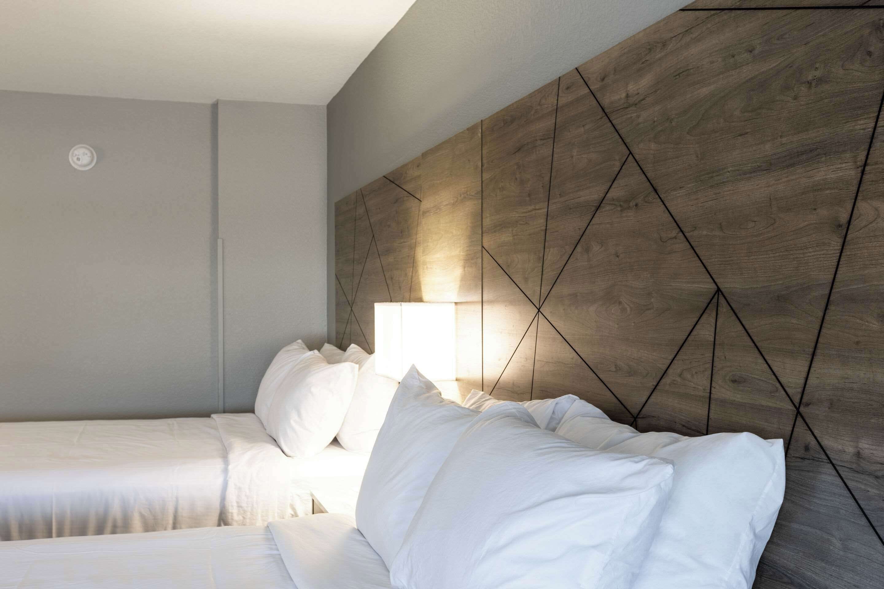 Habitació d'hotel amb dos llits, un panell de fusta al capçal i una tauleta de nit amb una làmpada.