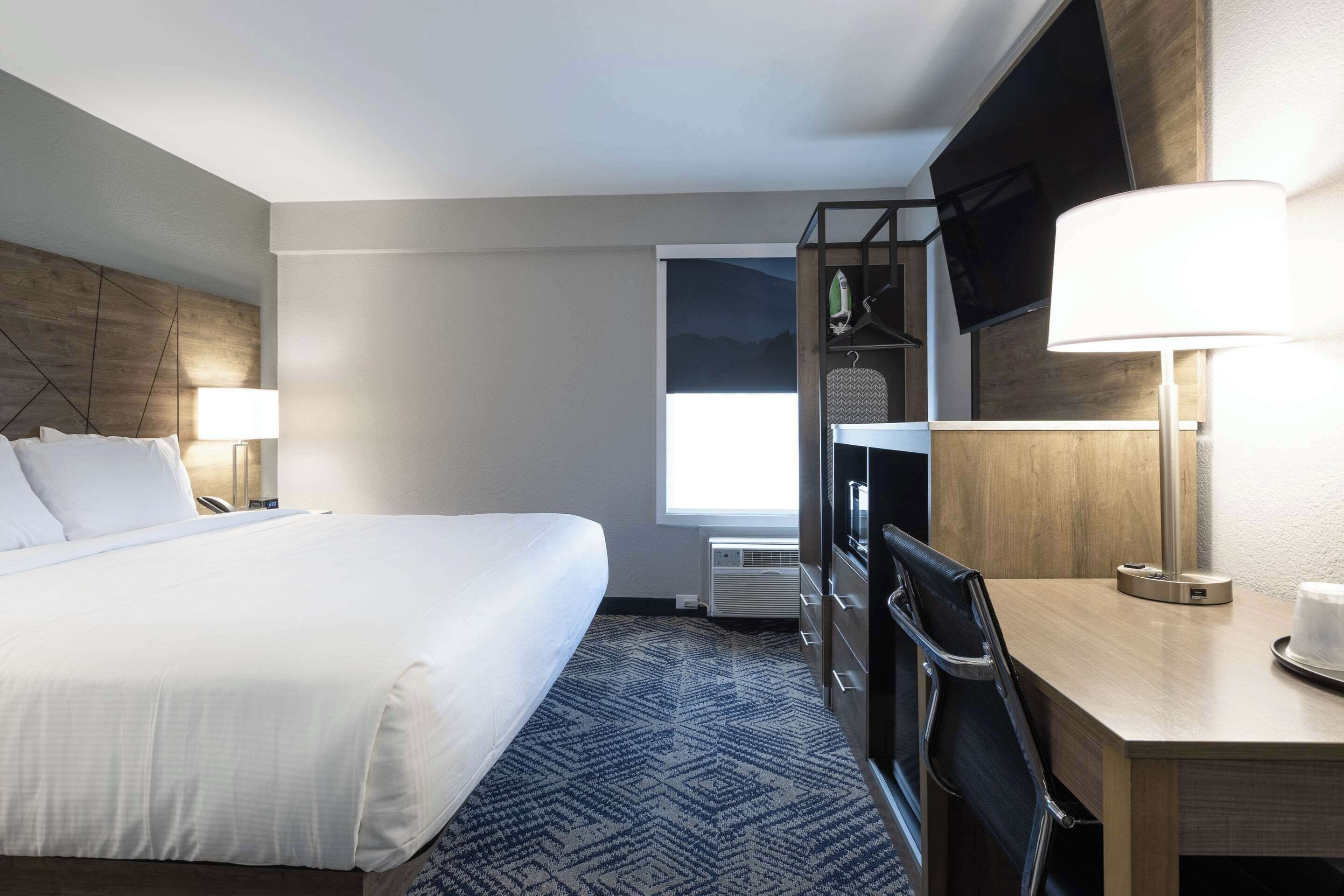 A hotel room with a large bed, a desk with a lamp, a TV mounted on the wall, and a window with a view of the outdoors.