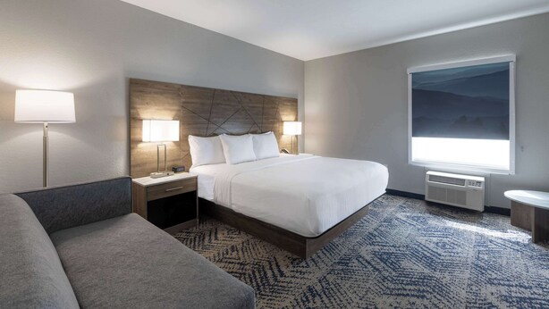 A hotel room with a large bed, two bedside lamps, a nightstand, a sofa, a large window with a view, and a wall-mounted air conditioner. - Rodeway Inn & Suites near Outlet Mall - Asheville (Asheville)