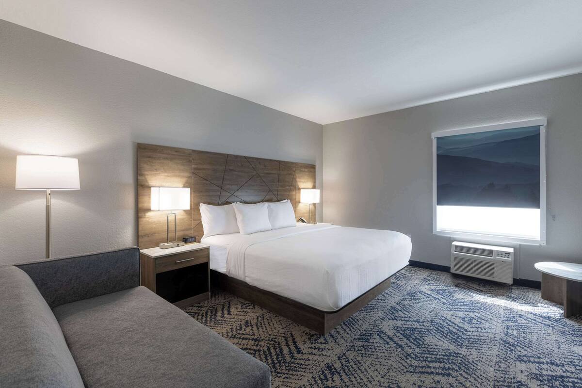 Quarto de hotel com uma cama grande, duas luminárias de cabeceira, uma mesa de cabeceira, um sofá, uma janela ampla com vista e um ar condicionado fixado na parede.