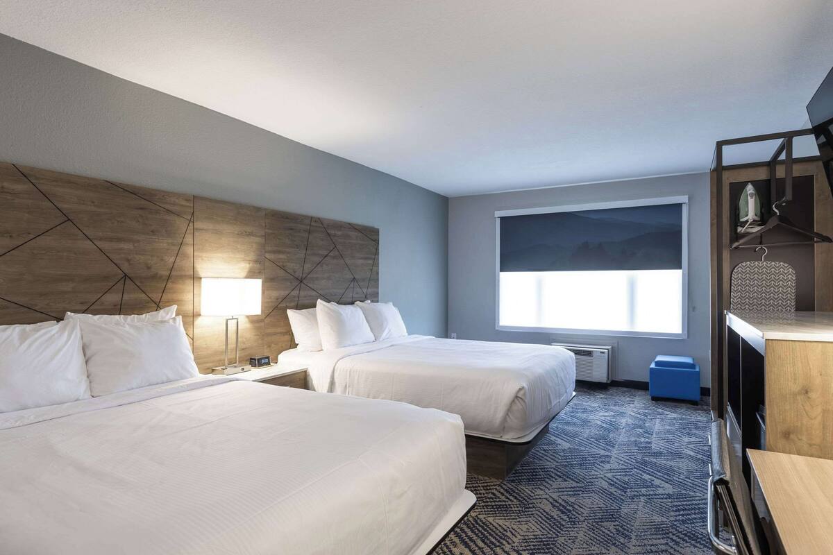 Quarto de hotel com duas camas, uma janela grande, uma escrivaninha com cadeira e um guarda-roupa.