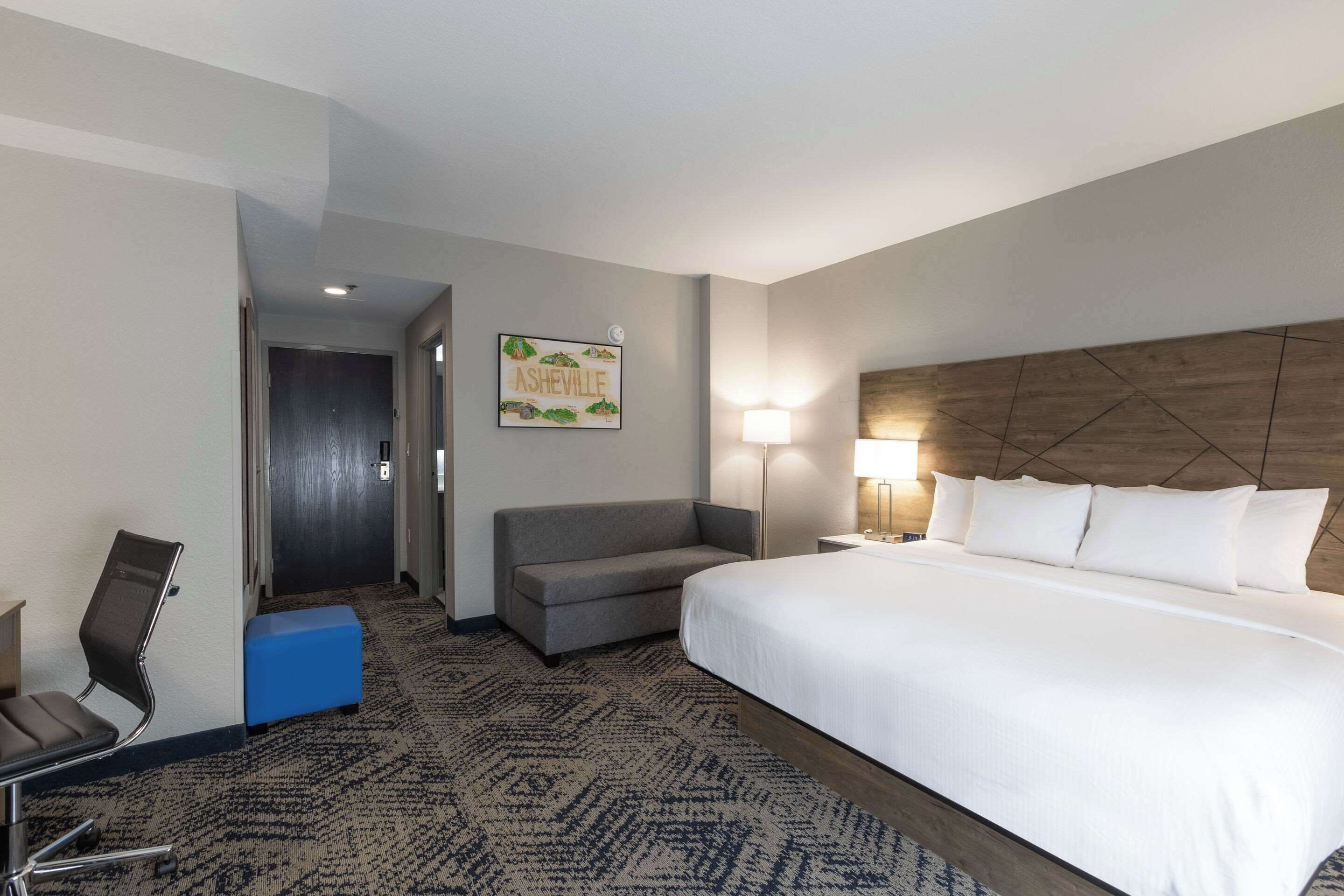 A hotel room with a large bed, a sofa, a desk with a chair, a lamp, and a wall-mounted artwork.