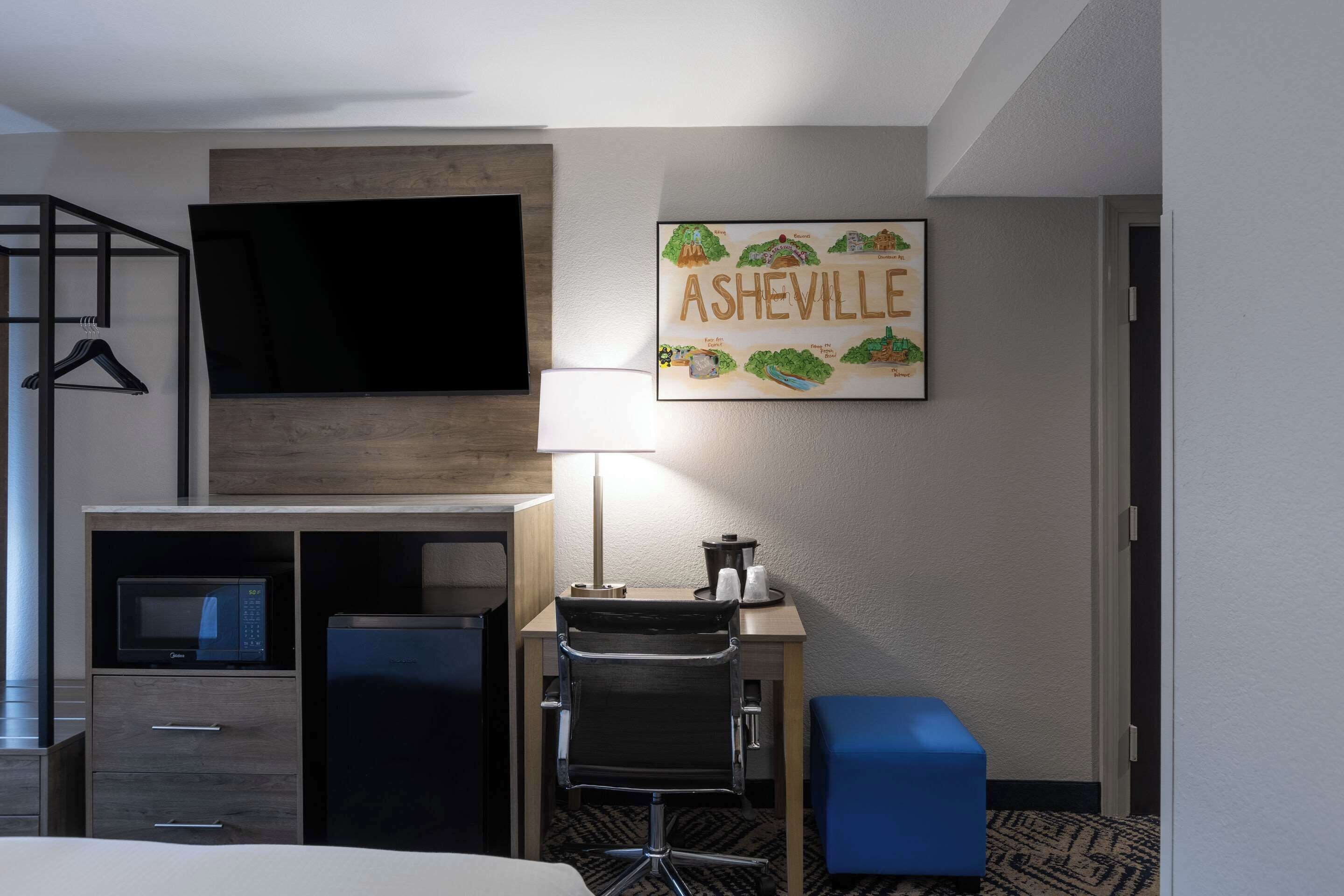 A hotel room with a bed, a desk with a chair, a TV, a microwave, a refrigerator, a lamp, and a framed picture of Asheville.