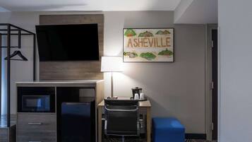 A hotel room with a bed, a desk with a chair, a TV, a microwave, a refrigerator, a lamp, and a framed picture of Asheville.