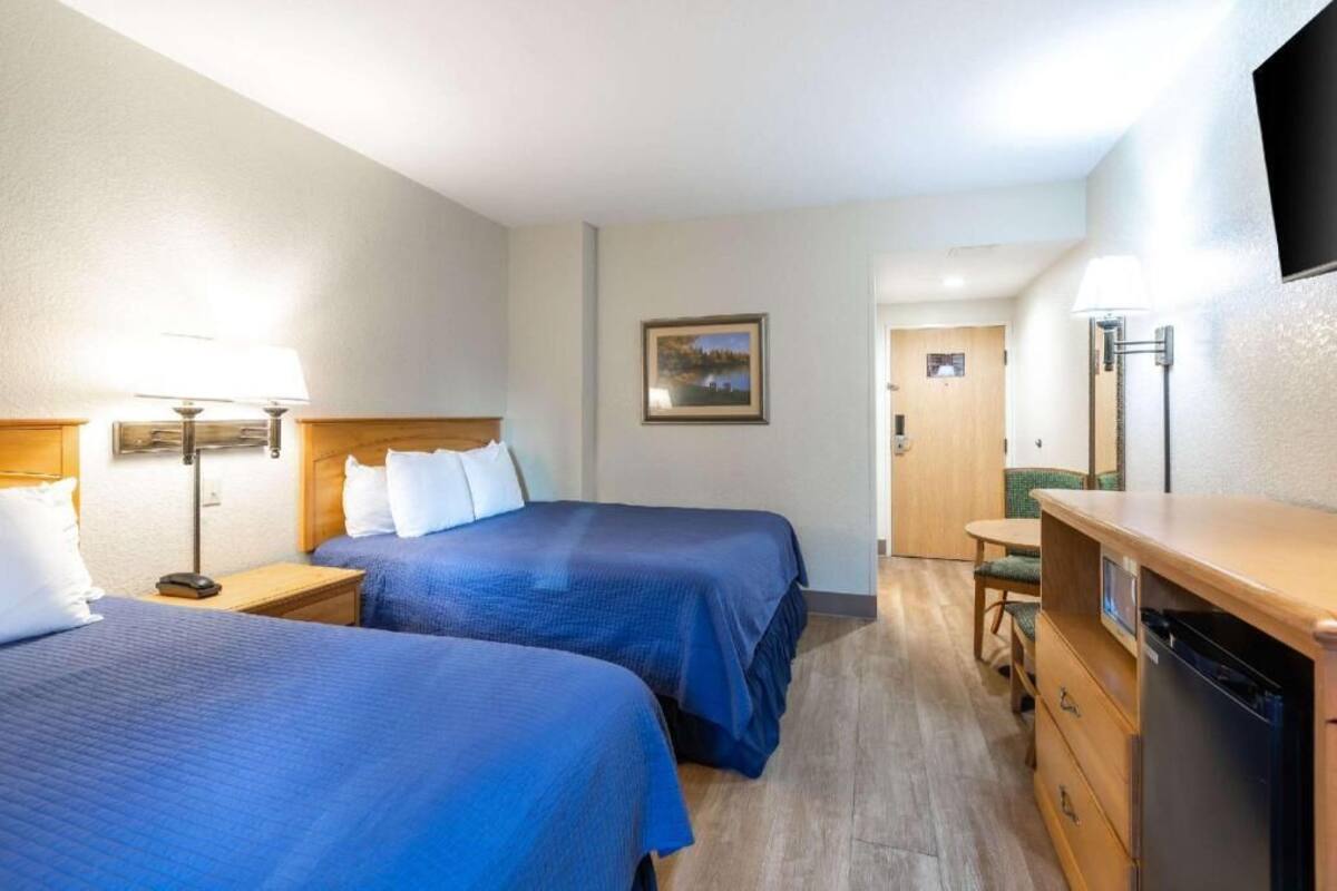 Quarto de hotel com duas camas, uma escrivaninha, uma cadeira, um frigorífico e uma porta que dá para outro espaço.