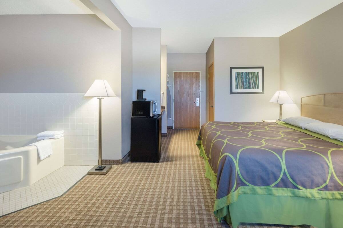 A hotel room with a bed, a desk with a lamp, a bathroom with a bathtub, and a door leading to another room.