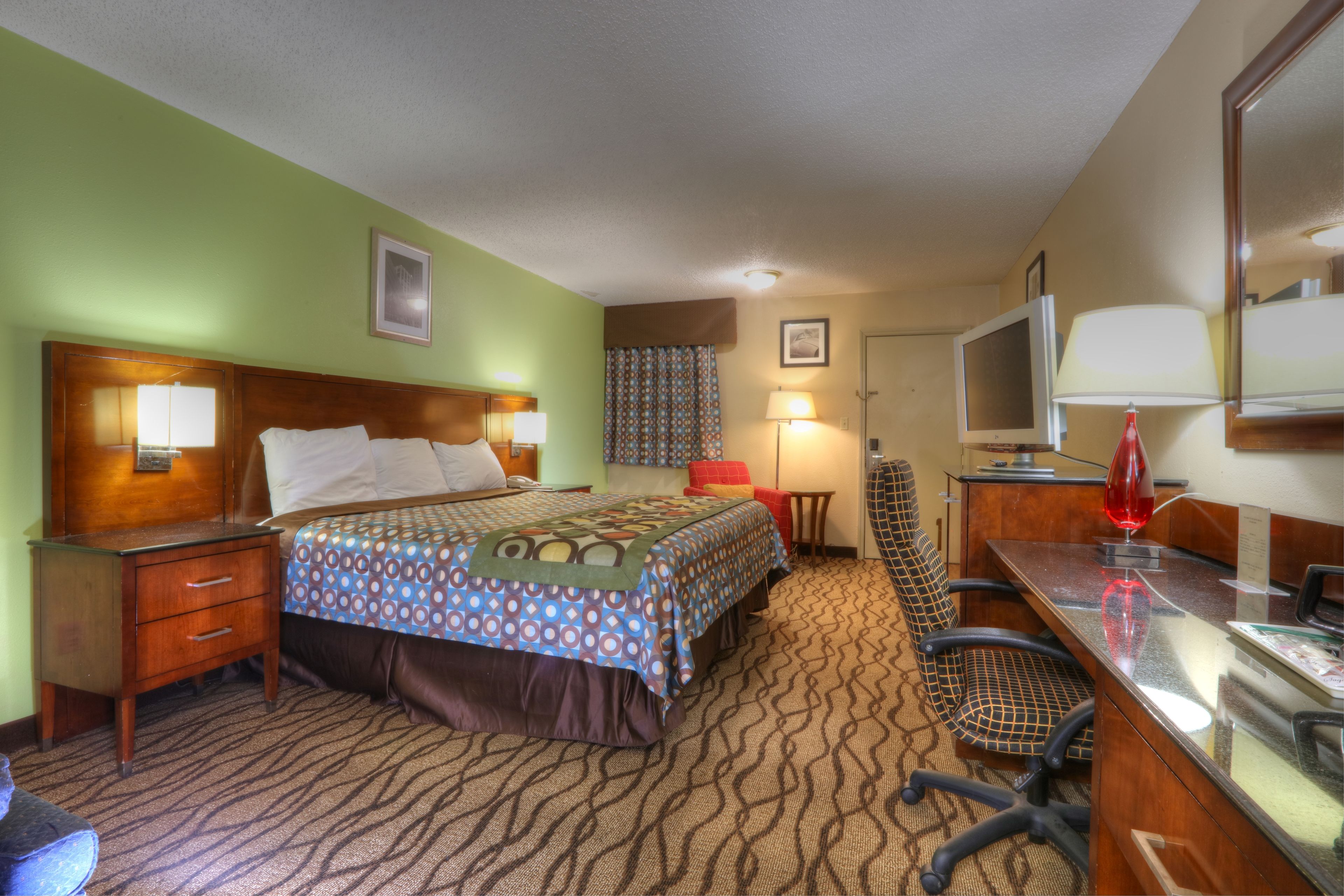 A hotel room with a bed, a desk with a computer, a chair, a TV, and a lamp.