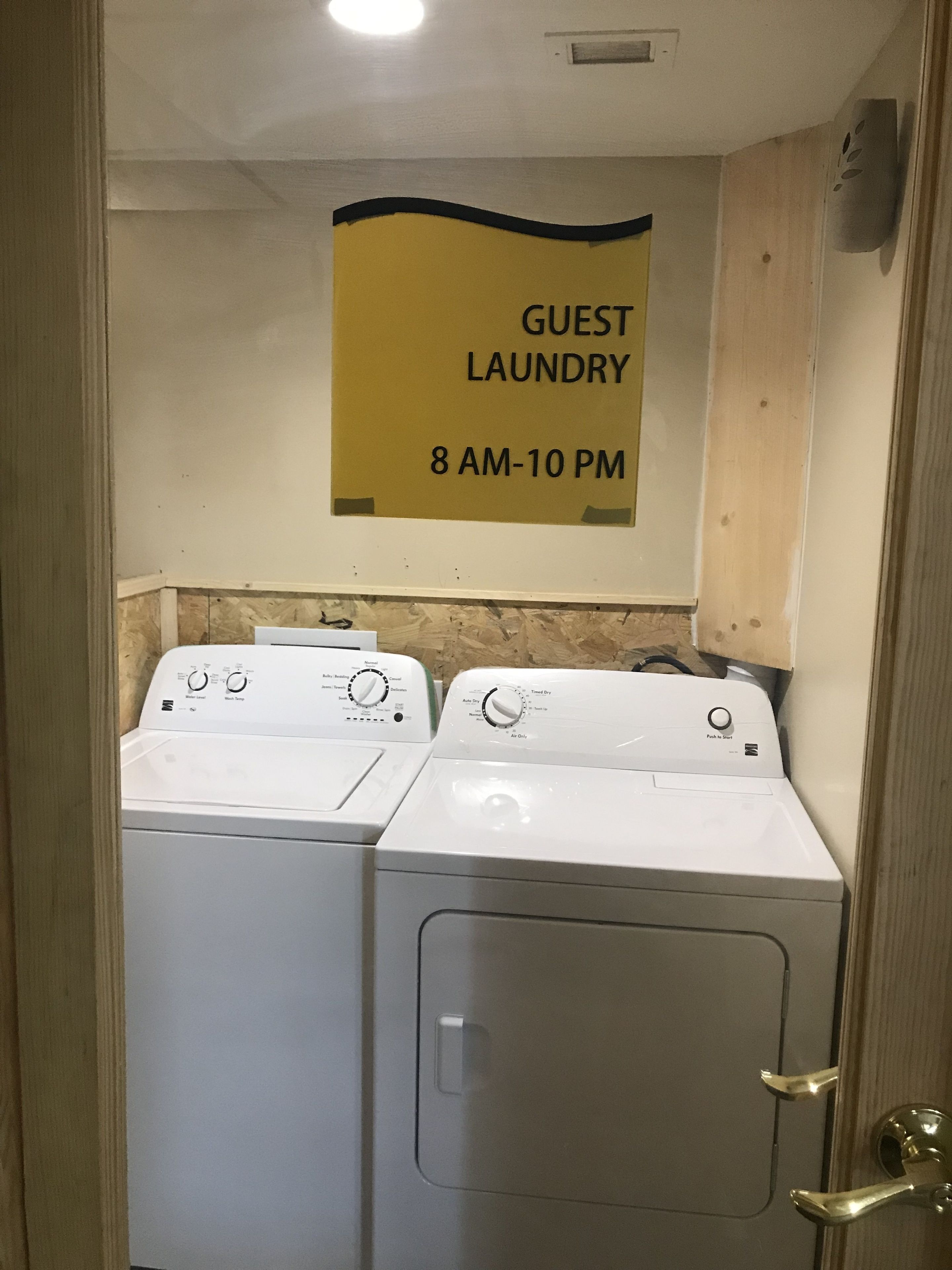 A laundry area with two washing machines, a sign indicating guest laundry hours, and a wooden wall.