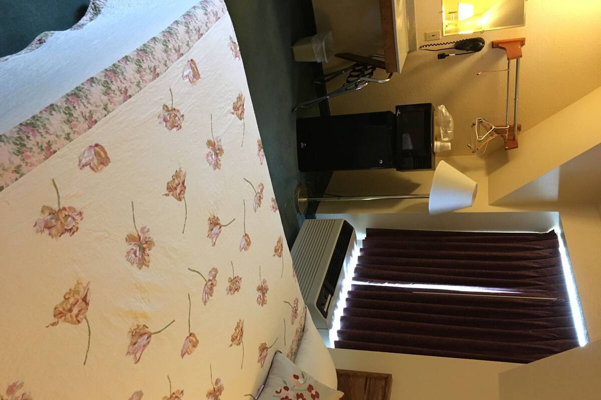 A bedroom with a floral bedspread, a heater, a window with curtains, and a wooden door.
