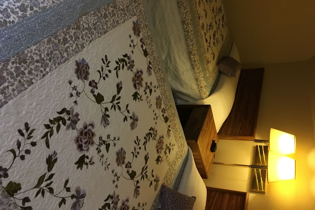 A bedroom with a floral bedspread, a bedside table with a lamp, and a wooden nightstand.