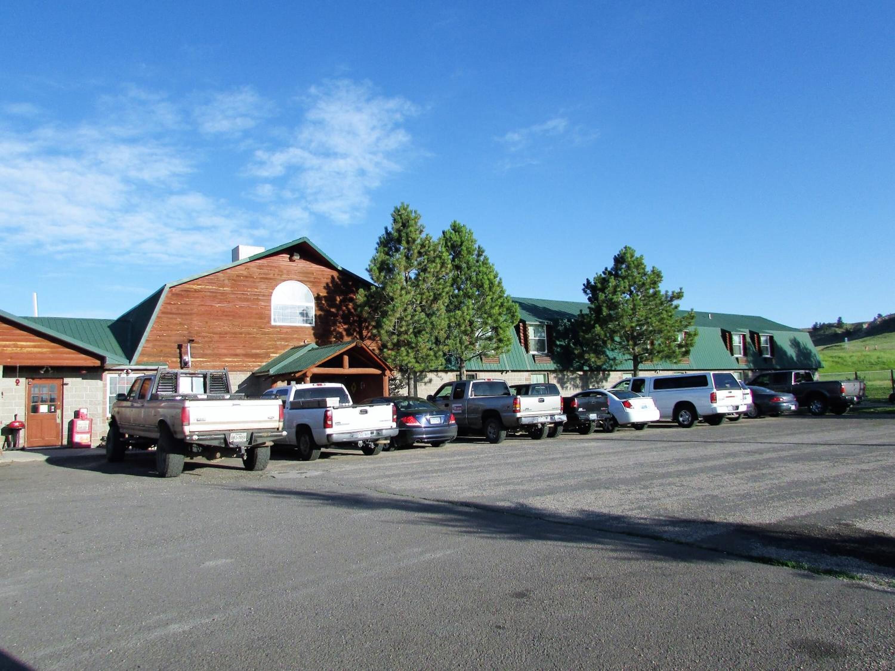 Un parking où sont garés plusieurs véhicules, dont des camions et des voitures, devant un grand bâtiment en bois surmonté d’un toit végétalisé.