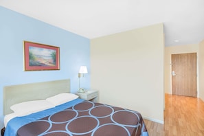 A hotel room with a bed, a nightstand, a lamp, a painting, and a wooden door.