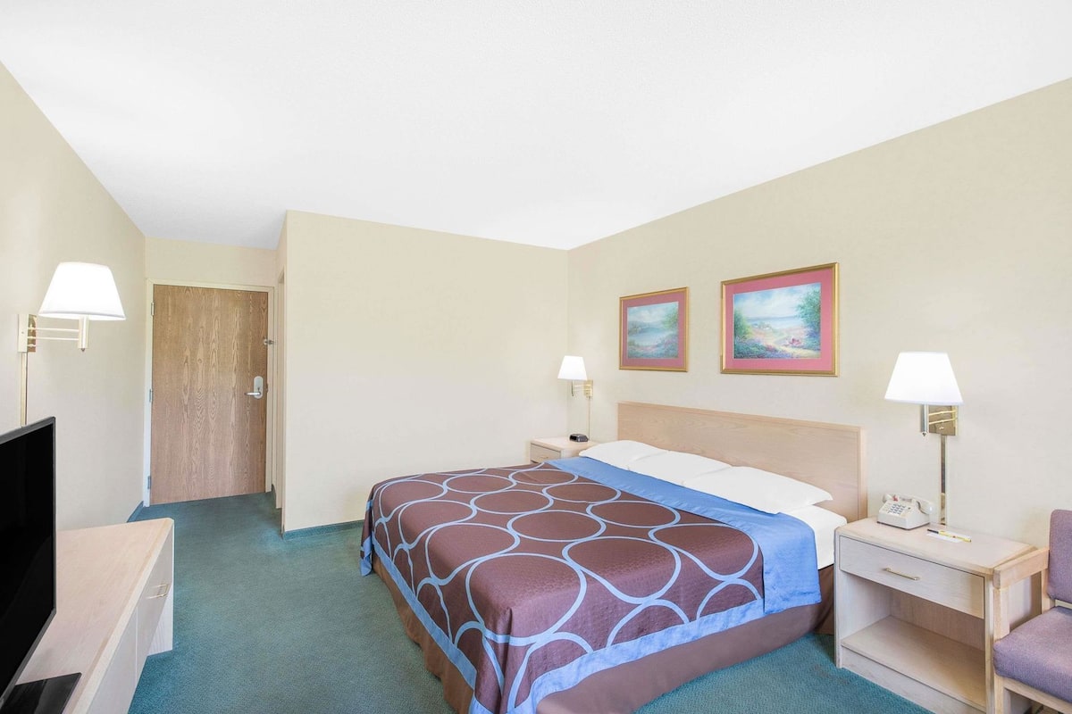 A hotel room with a bed, two bedside tables, a desk, a chair, a TV, and two paintings on the wall.