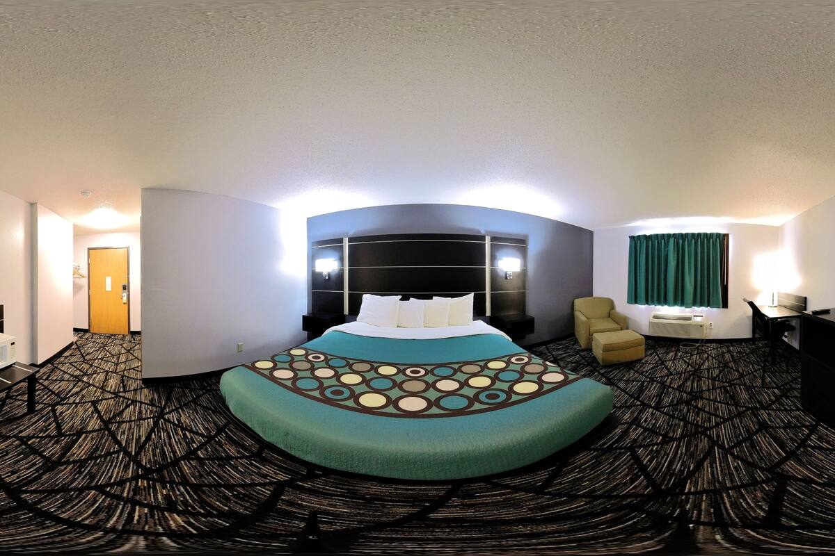 A hotel room with a curved bed, a headboard with lights, a desk, a chair, and a window with curtains.