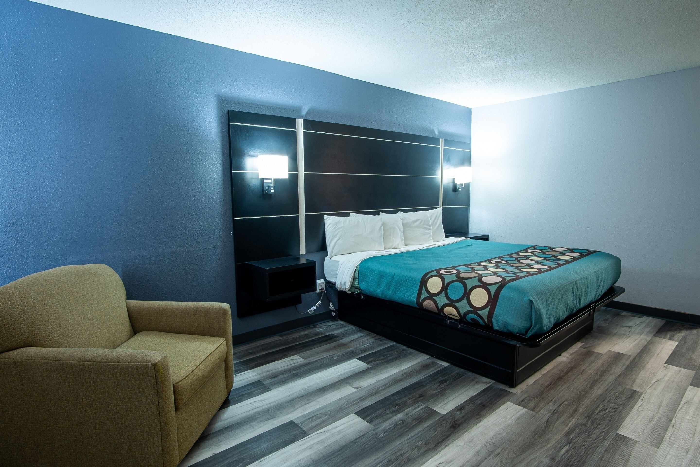 A modern hotel room with a bed, a nightstand, a chair, and a wall-mounted light fixture.