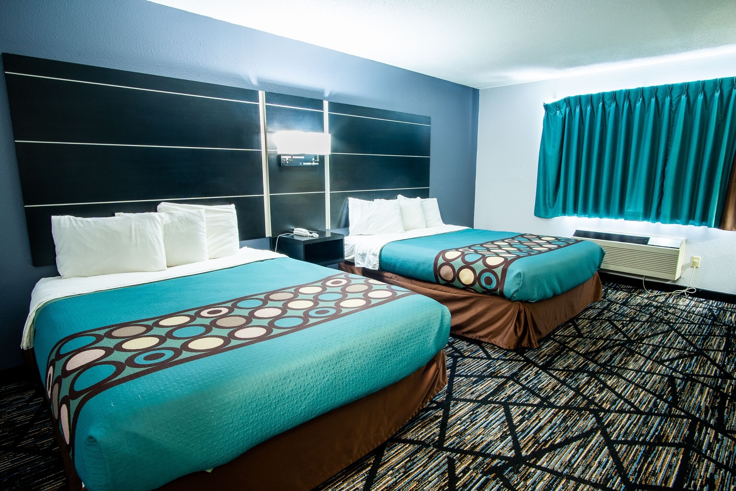 A hotel room with two beds, a large headboard, a window with curtains, and a patterned carpet.