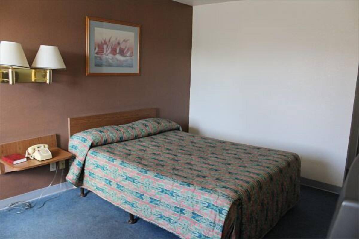 A hotel room with a single bed, a bedside table with a lamp, a phone, and a framed picture on the wall.