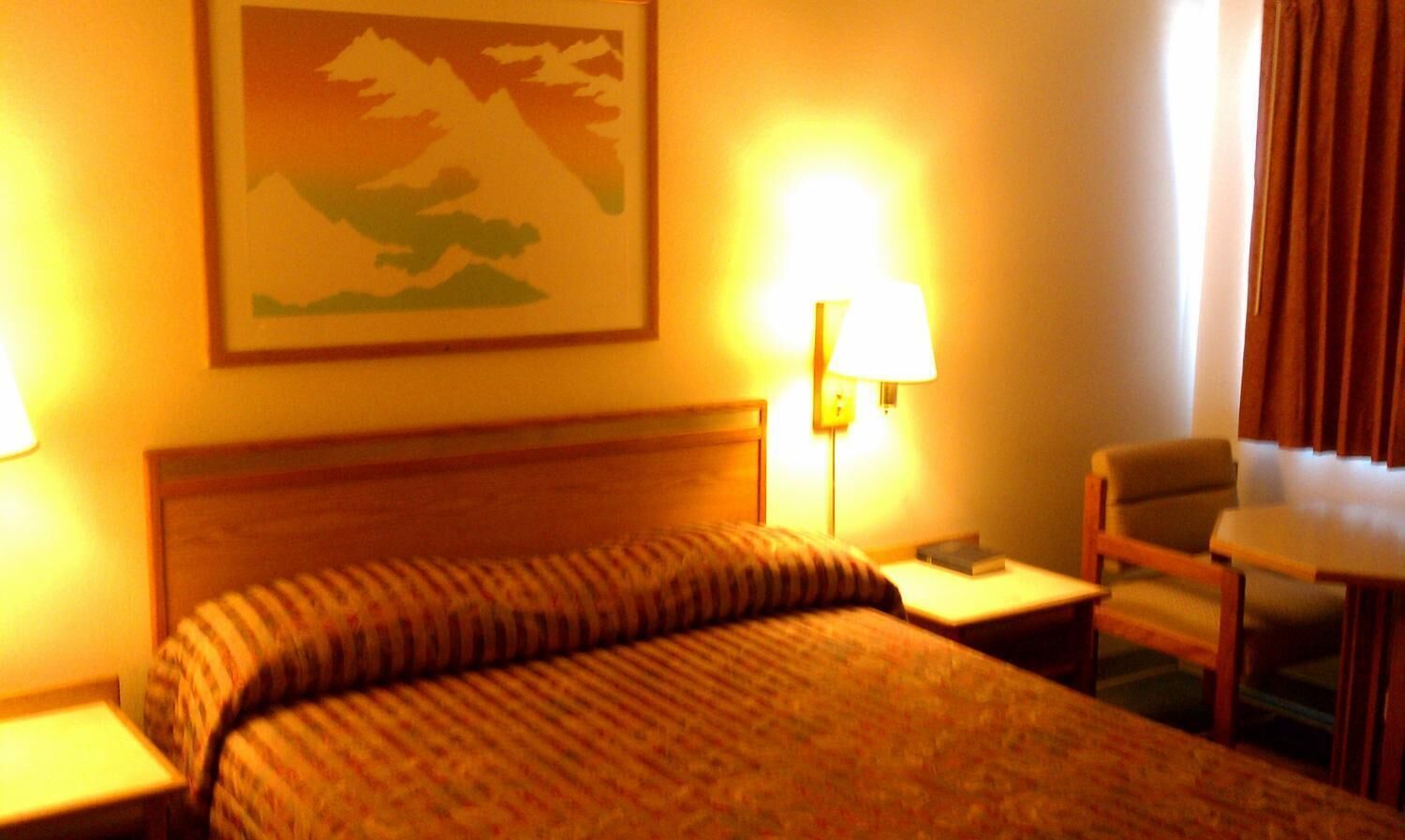 A hotel room with a bed, two bedside lamps, a chair, a small table, and a painting on the wall.
