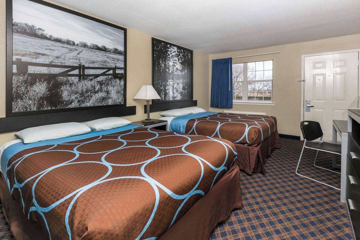 Habitación de hotel con dos camas, un escritorio, una silla, un ventanal grande con cortinas y un cuadro en la pared.