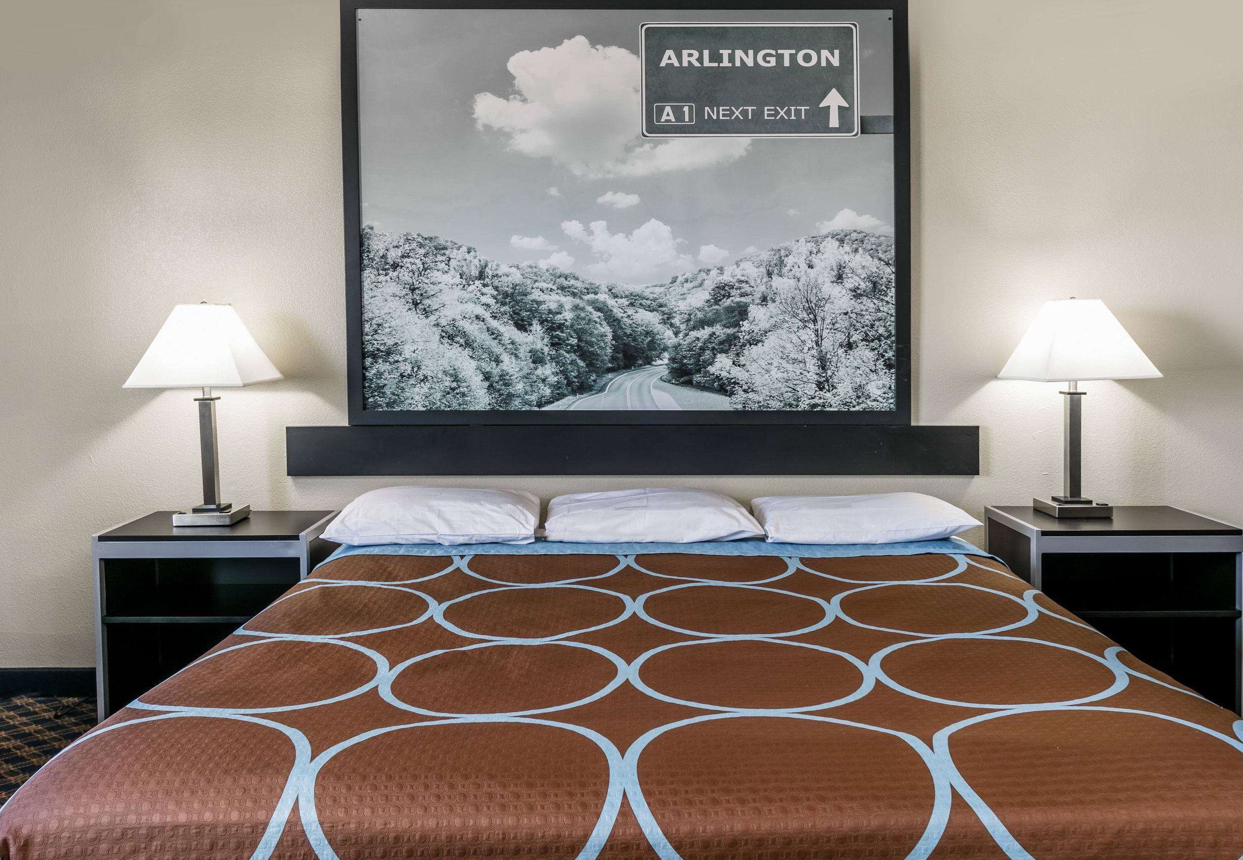 A hotel room with a bed, two bedside tables with lamps, a large framed picture of a road, and a sign indicating the next exit to Arlington.