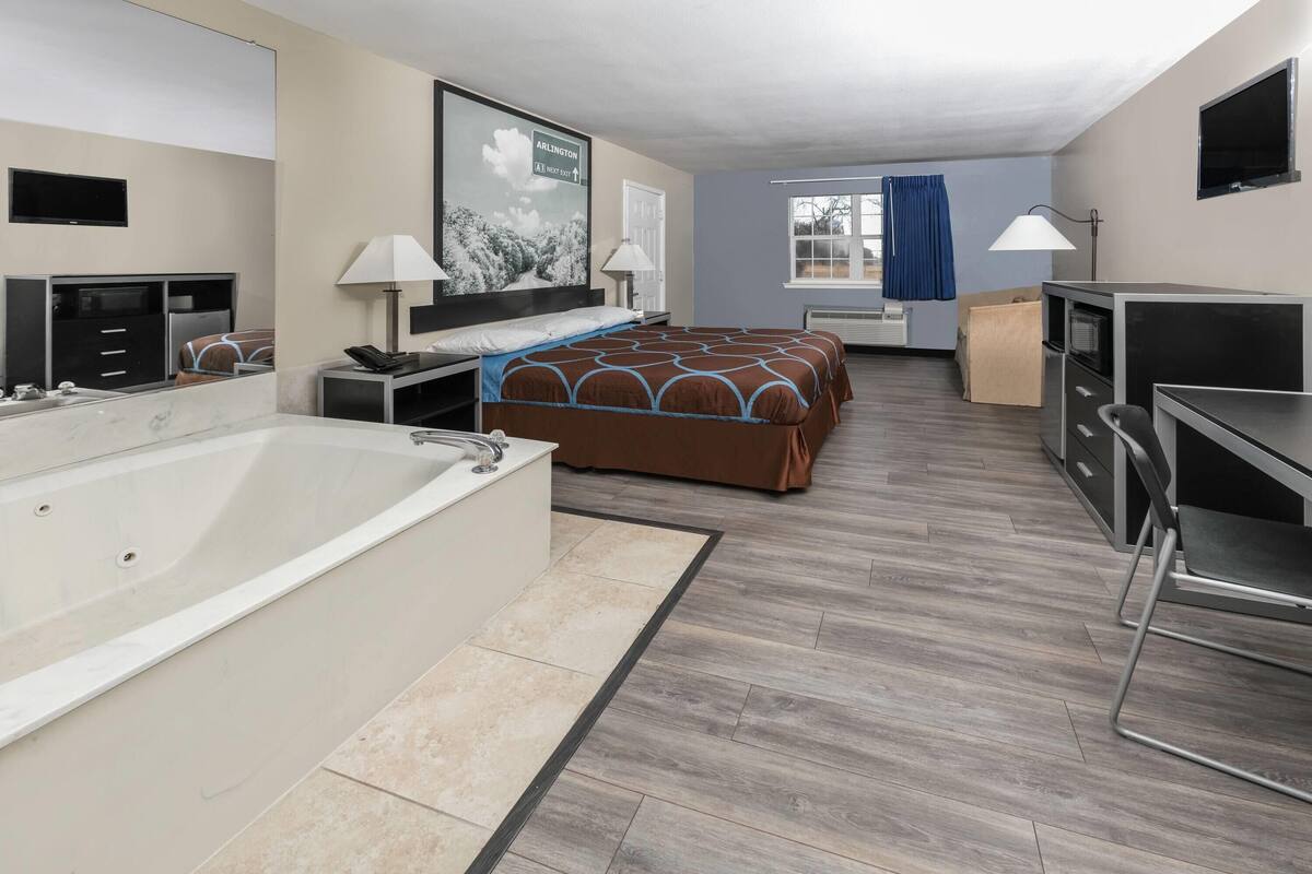 Habitación de hotel con una cama grande, un escritorio independiente con silla, bañera, televisor montado en la pared y ventana con cortinas.