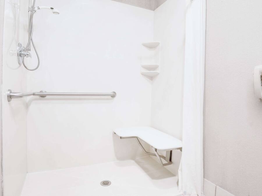 A white bathroom with a shower, a handrail, a bench, and a wall-mounted phone.
