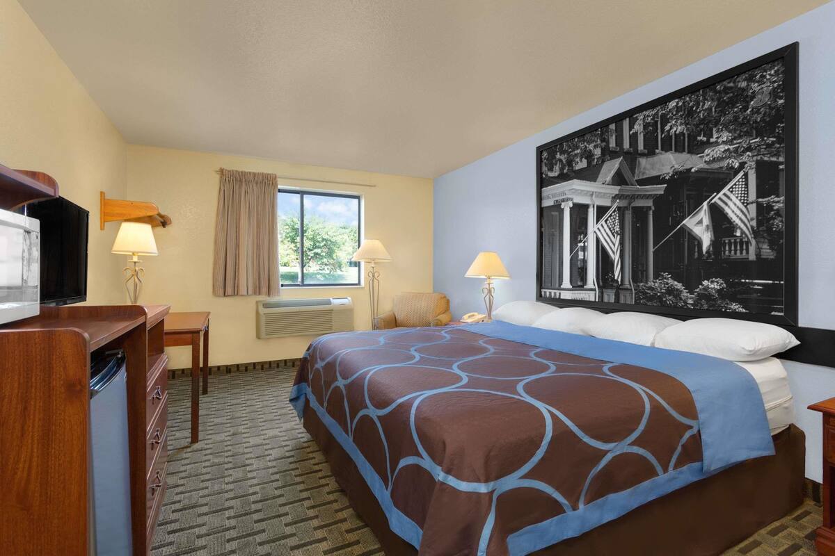A hotel room with a bed, a desk, a television, two lamps, a window with curtains, and a large framed picture on the wall.