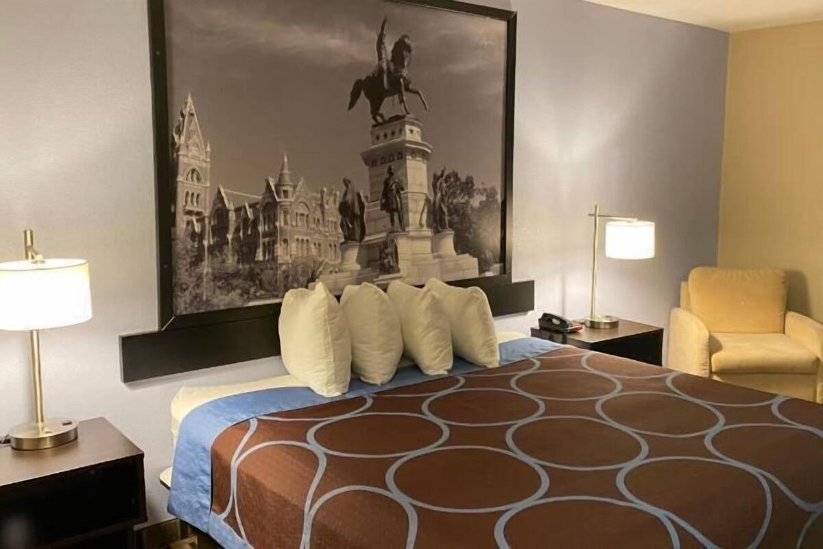 A hotel room with a large bed, a chair, a nightstand, a lamp, and a large framed picture on the wall.