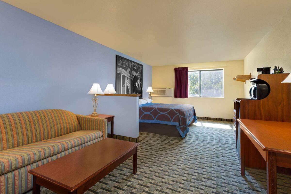 A hotel room with a striped sofa, a wooden table, a bed with a blue patterned bedspread, a window with a view, and a wall-mounted air conditioning unit.