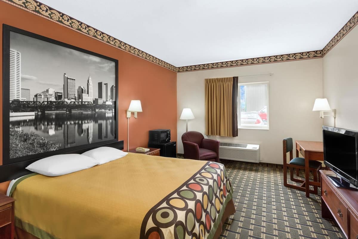 A hotel room with a bed, a desk, a chair, a television, a window with curtains, a wall-mounted lamp, and a large cityscape painting.