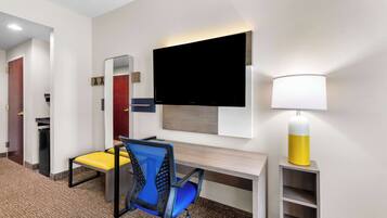 A hotel room with a desk, a chair, a TV mounted on the wall, a lamp, and a mirror.