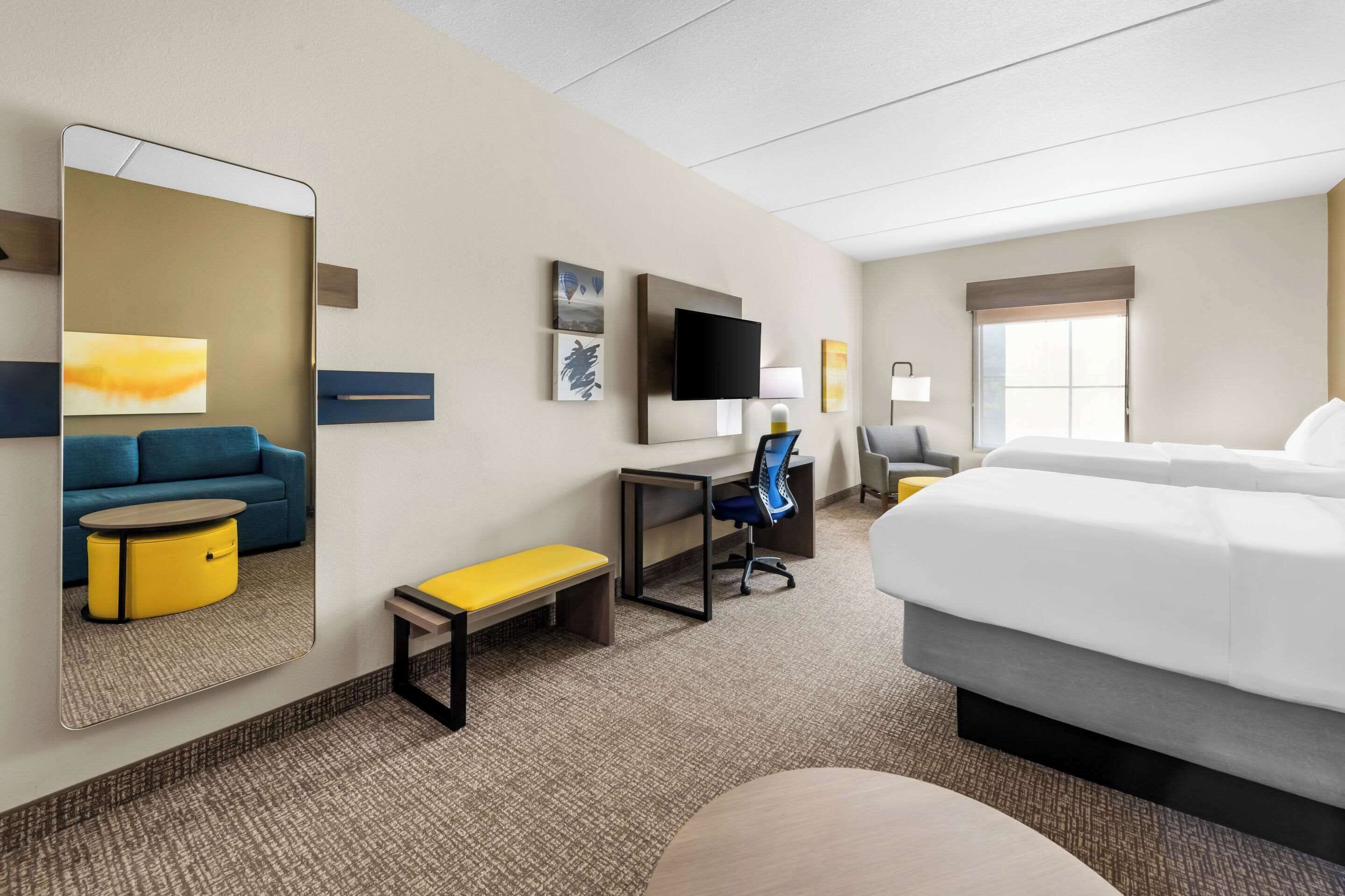 A hotel room with a large bed, a desk with a chair, a sofa, a small table, a bench, and a TV on the wall.