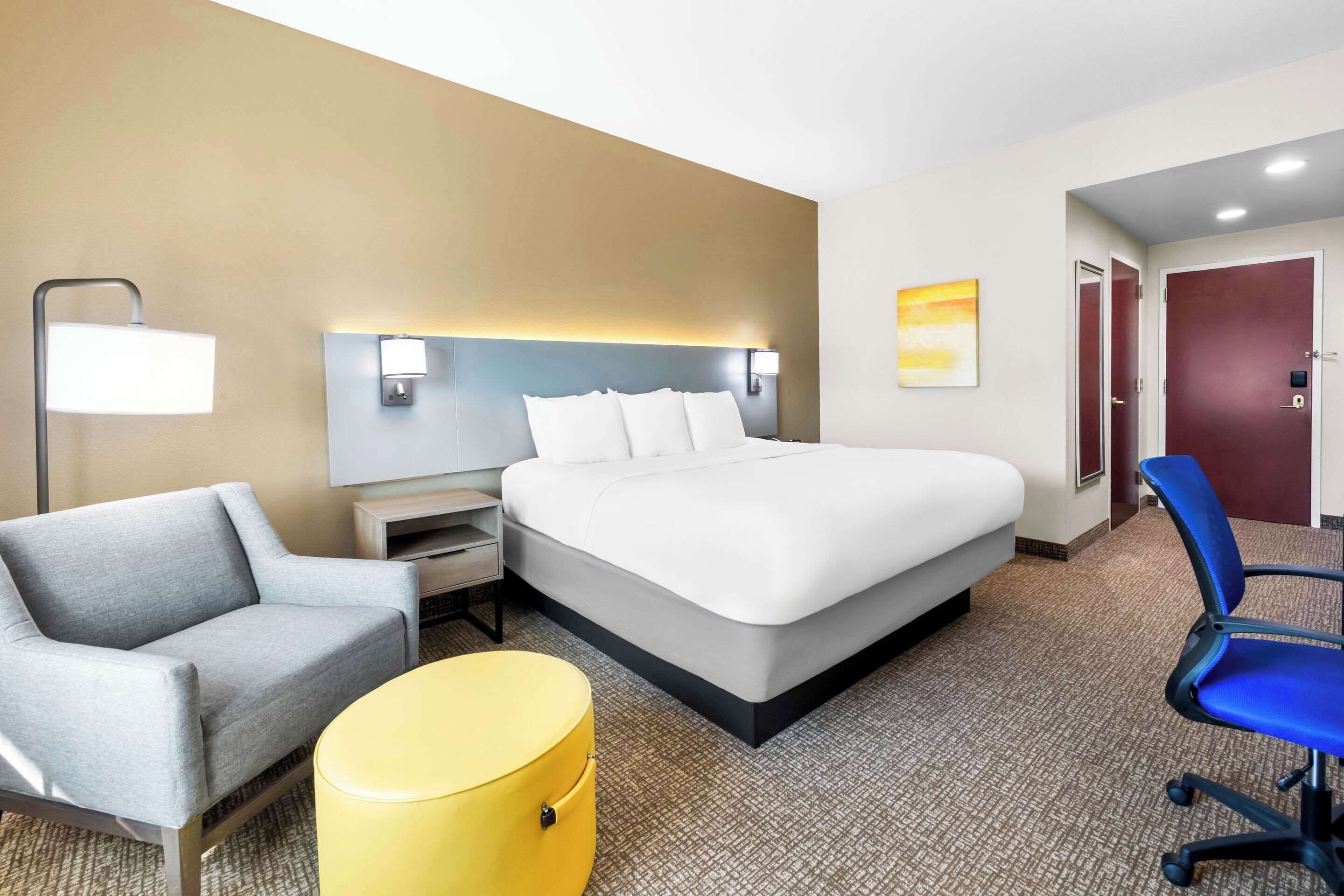 A hotel room with a bed, a gray armchair, a yellow ottoman, a blue office chair, a nightstand, and a wall-mounted lamp.