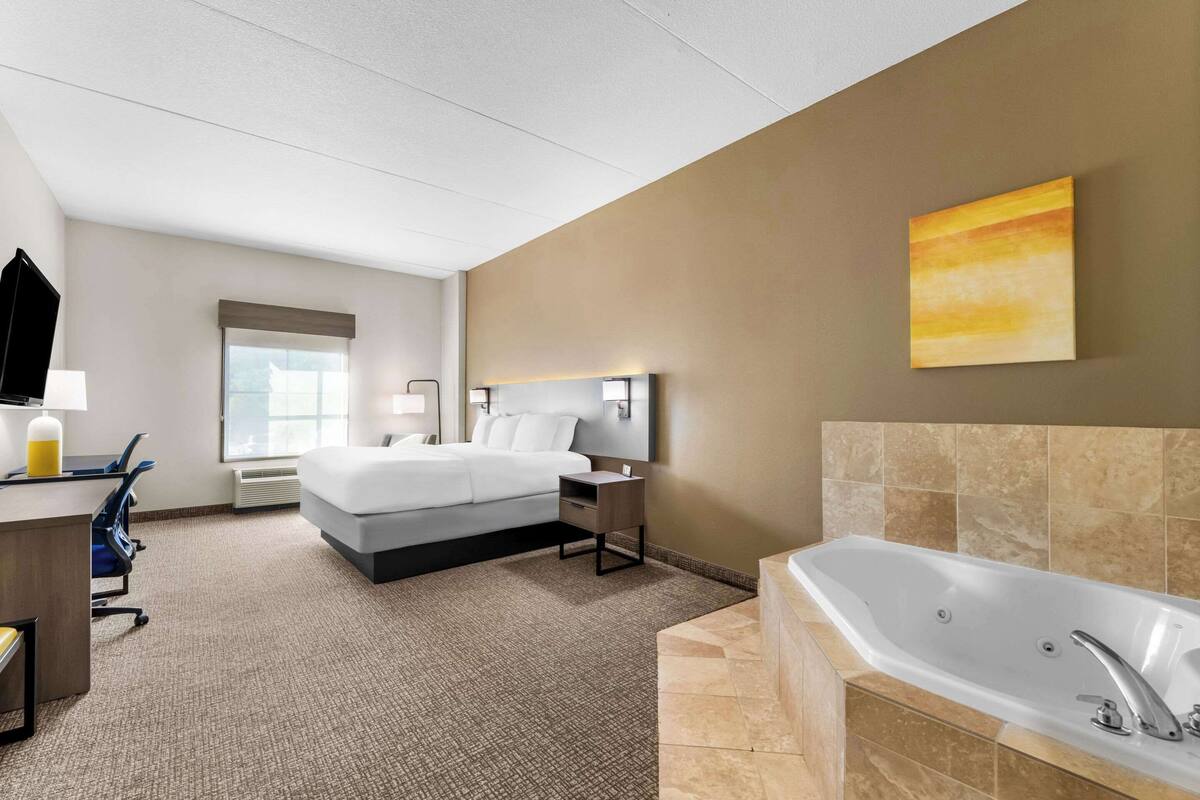 A hotel room with a bed, a desk with a chair, a desk lamp, a wall-mounted TV, a large window, a bathroom with a bathtub, and a painting on the wall.