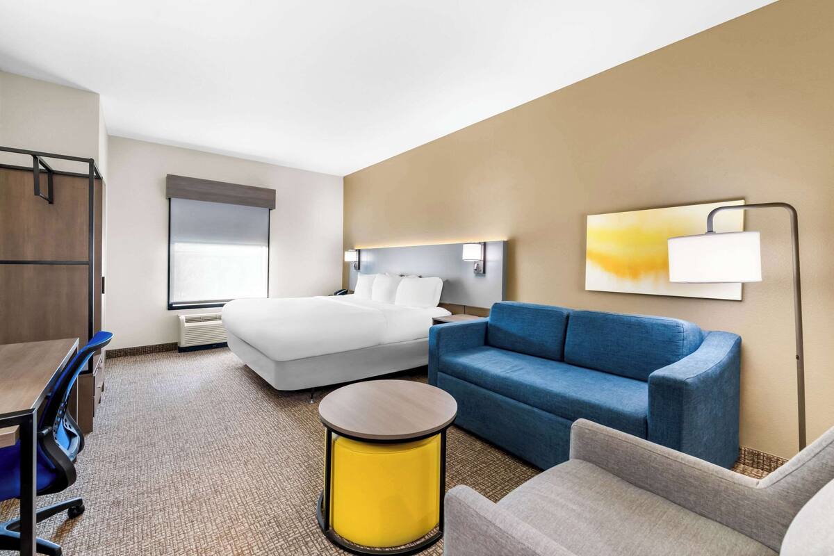 A hotel room with a bed, a blue sofa, a small round table, a desk with a chair, and a large window.