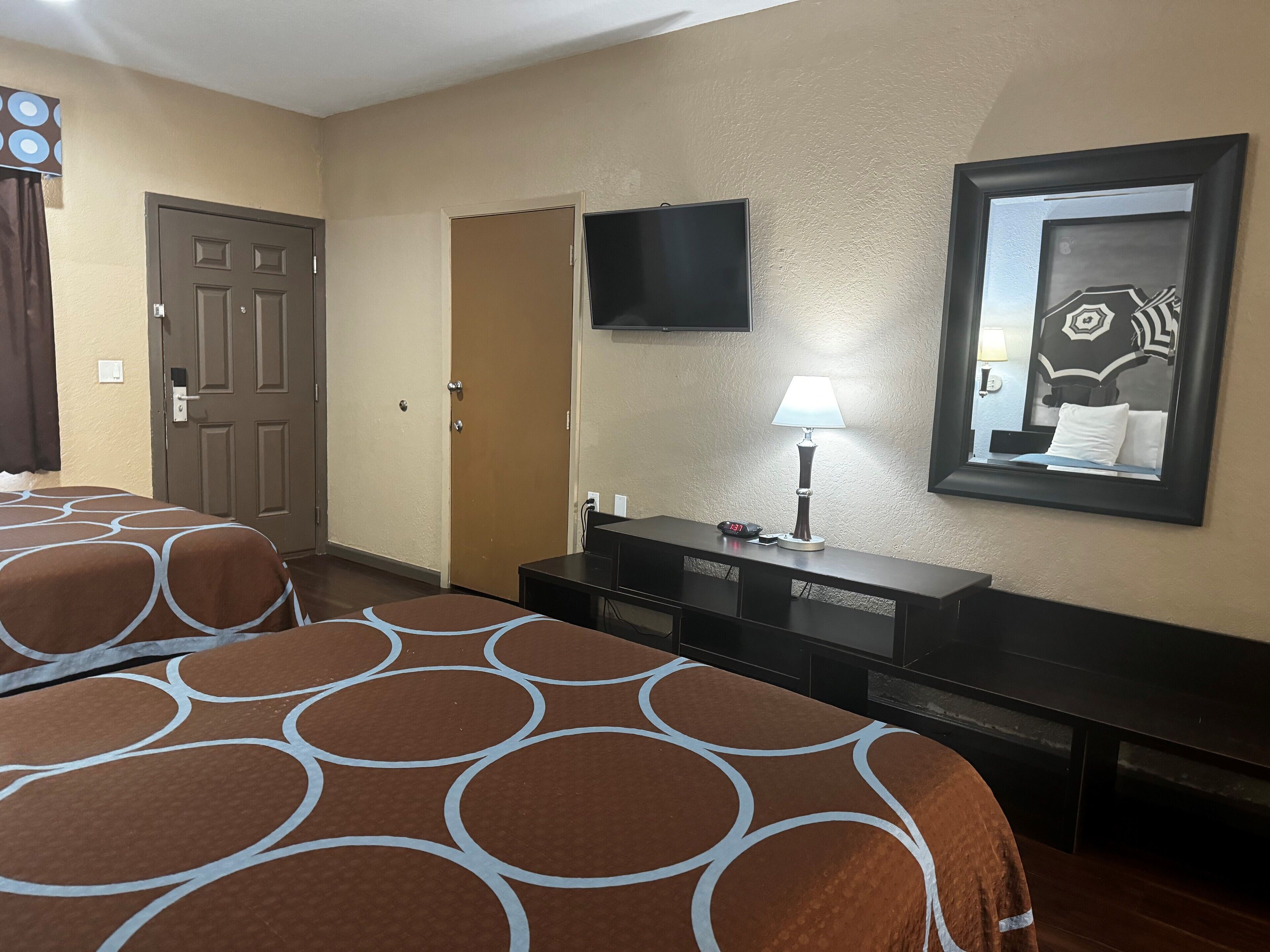 Habitación de hotel con dos camas, un televisor montado en la pared, un escritorio con una lámpara y un espejo que refleja parte de la habitación.