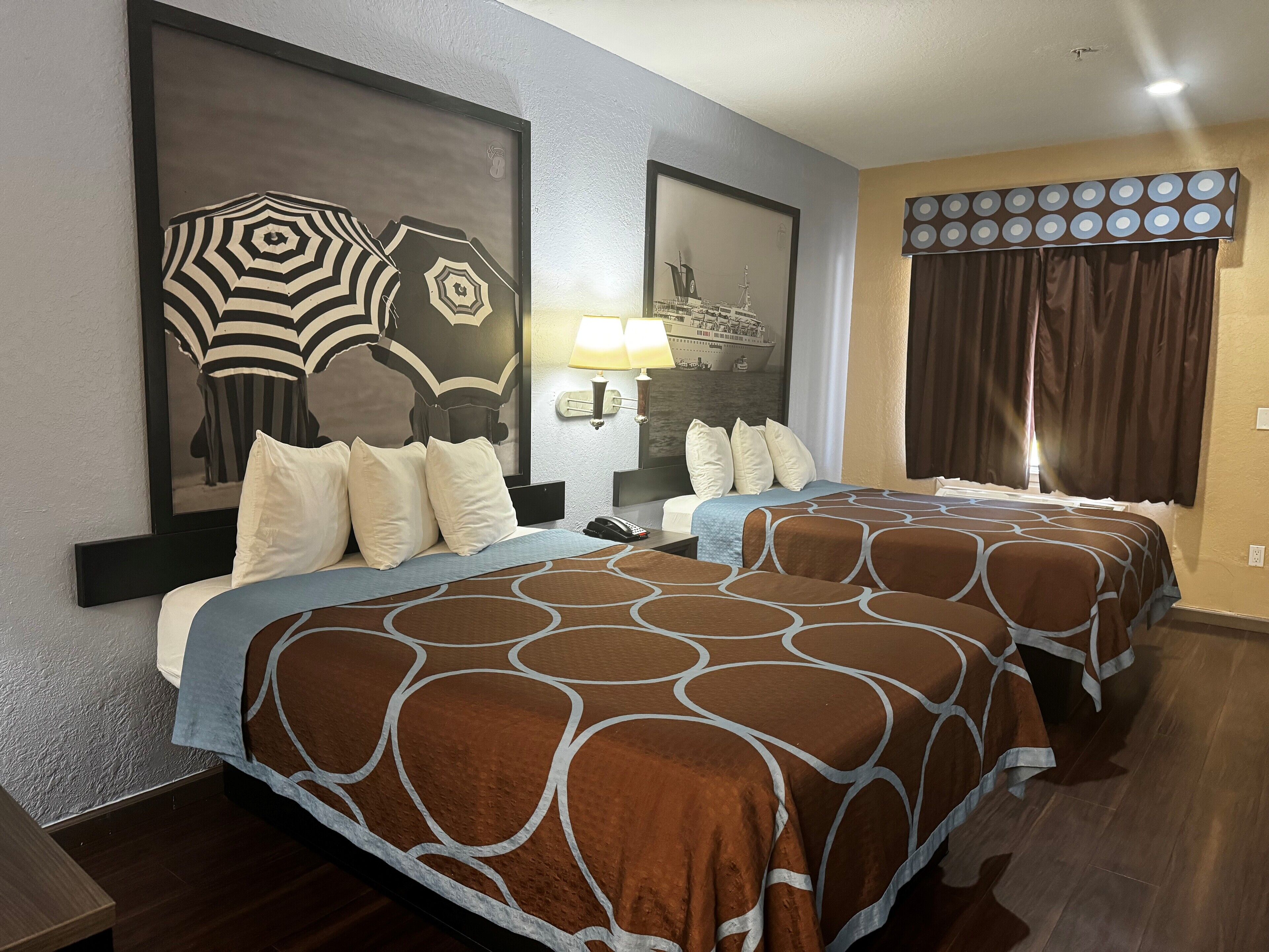 A hotel room with two beds, a large framed artwork above the left bed, a nightstand with a lamp, and a window with curtains.
