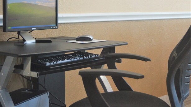 Ein Schreibtisch mit Monitor, Tastatur und Maus. An der Wand hängen zwei gerahmte Bilder.
