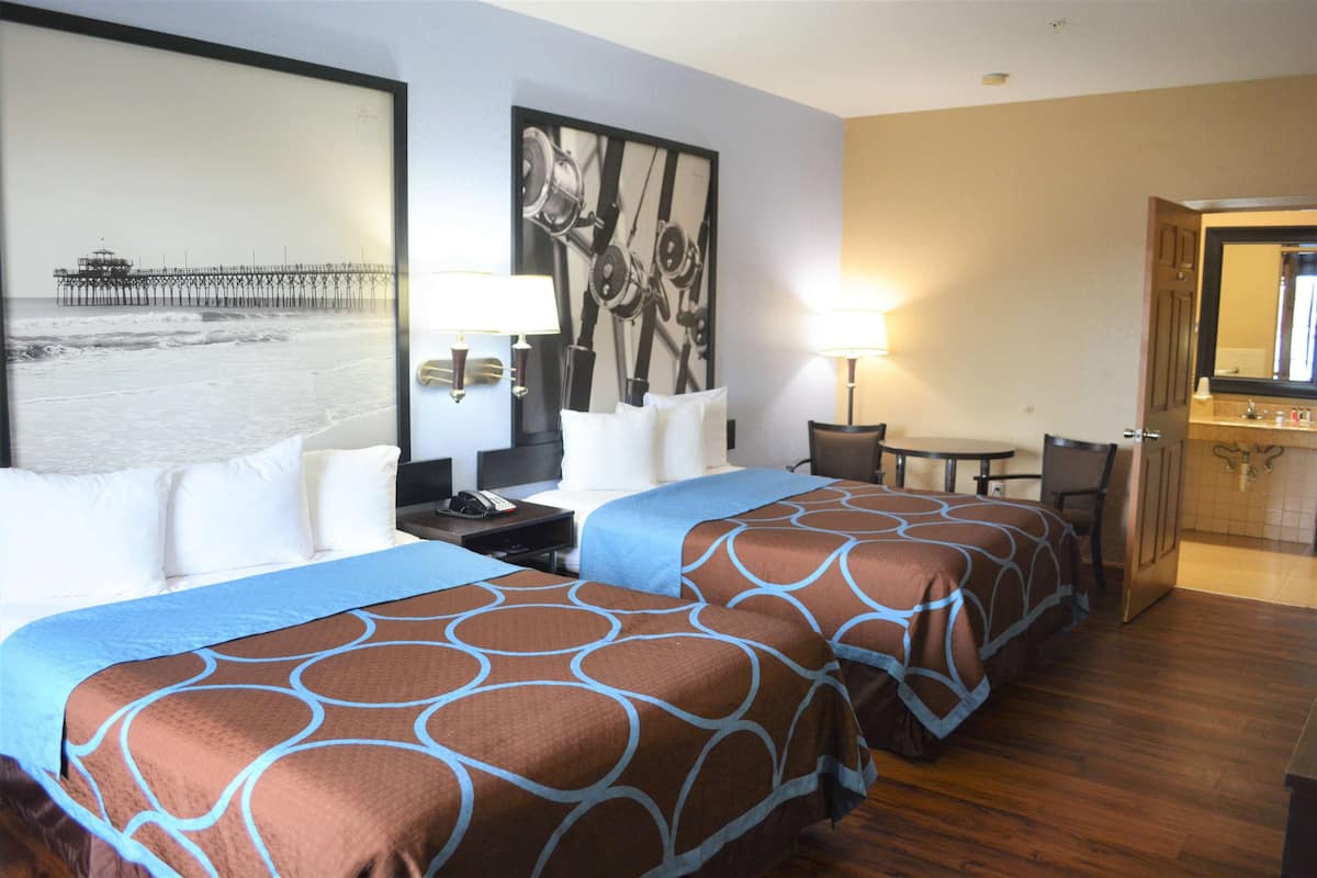 Ein Hotelzimmer mit zwei Betten, einem Schreibtisch, einem Stuhl, einem kleinen Tisch und einem durchgegangene Tür erkennbaren Badezimmer.