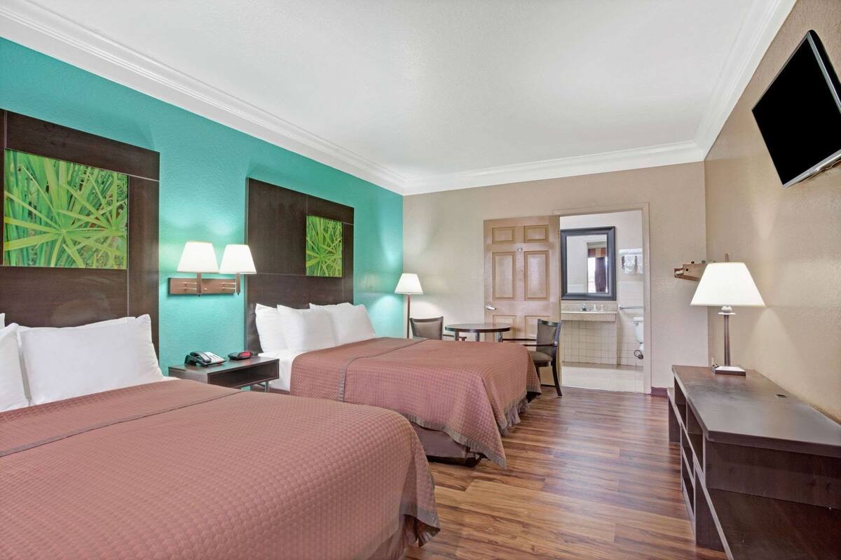Ein Hotelzimmer mit zwei Betten, einem Schreibtisch, einem Stuhl, einem kleinen Tisch, einem Fernseher und einem durchgegangenen Badezimmer.