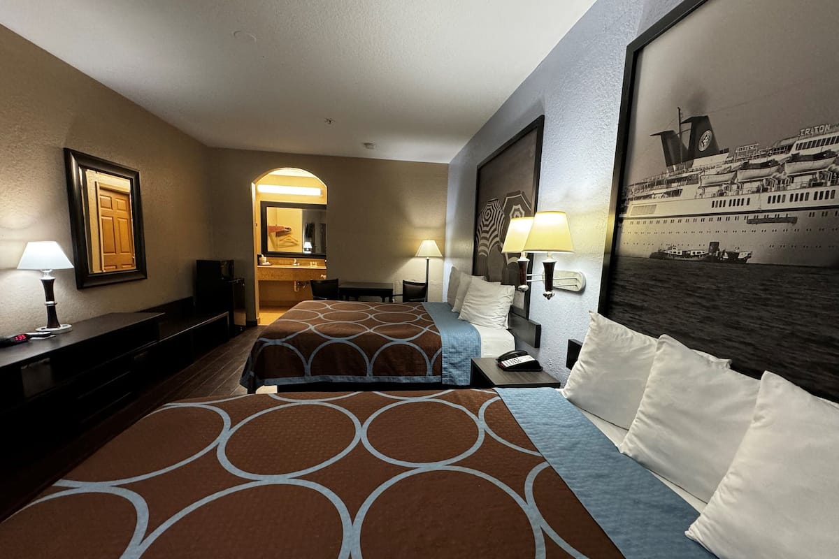 Ein Hotelzimmer mit zwei Betten, einem Schreibtisch, einem Stuhl, einem Spiegel und einem an der Wand befestigten Fernseher.