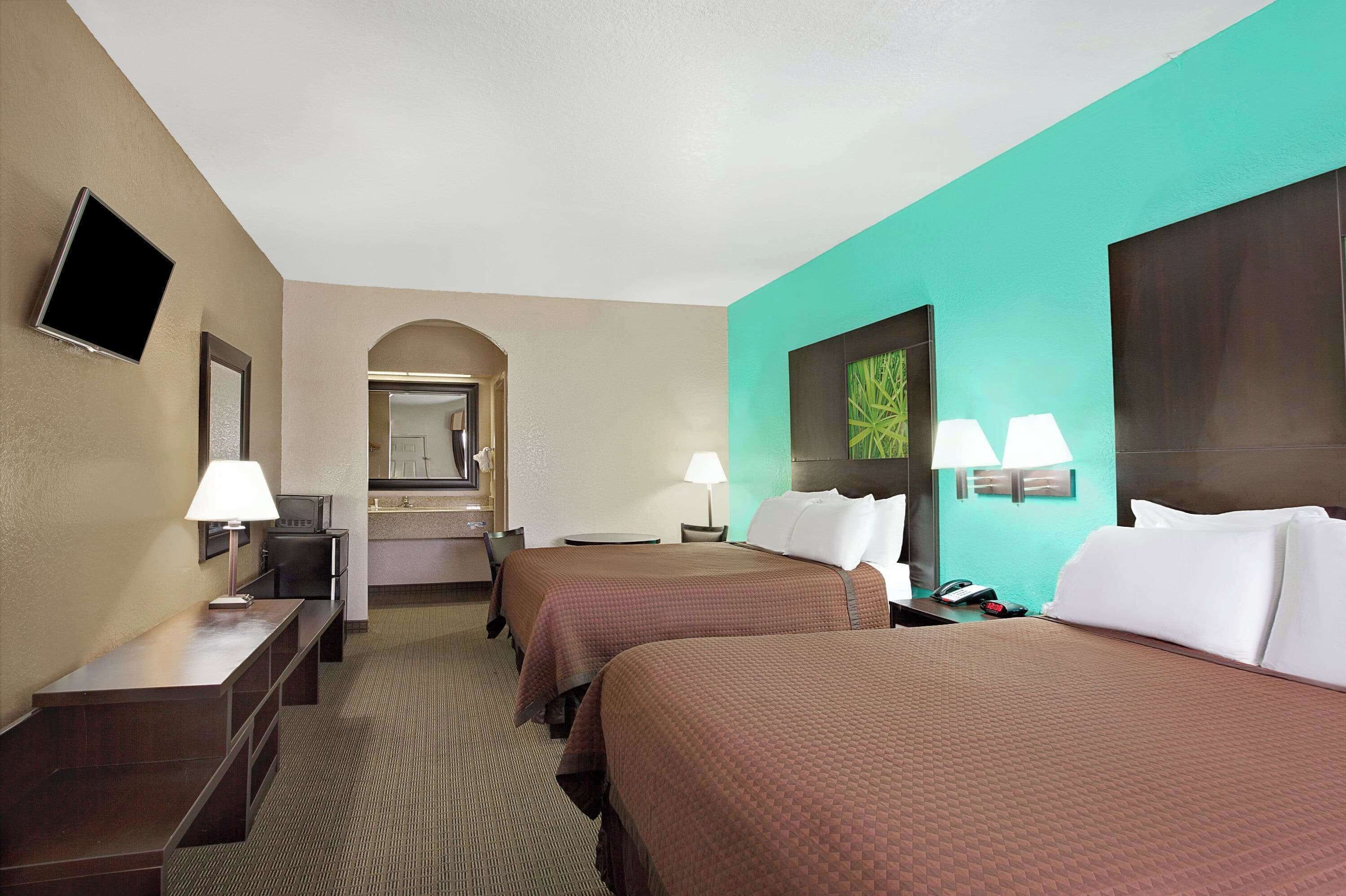 Habitación de hotel con dos camas, un escritorio, una silla, un televisor y un baño visible a través de un arco.