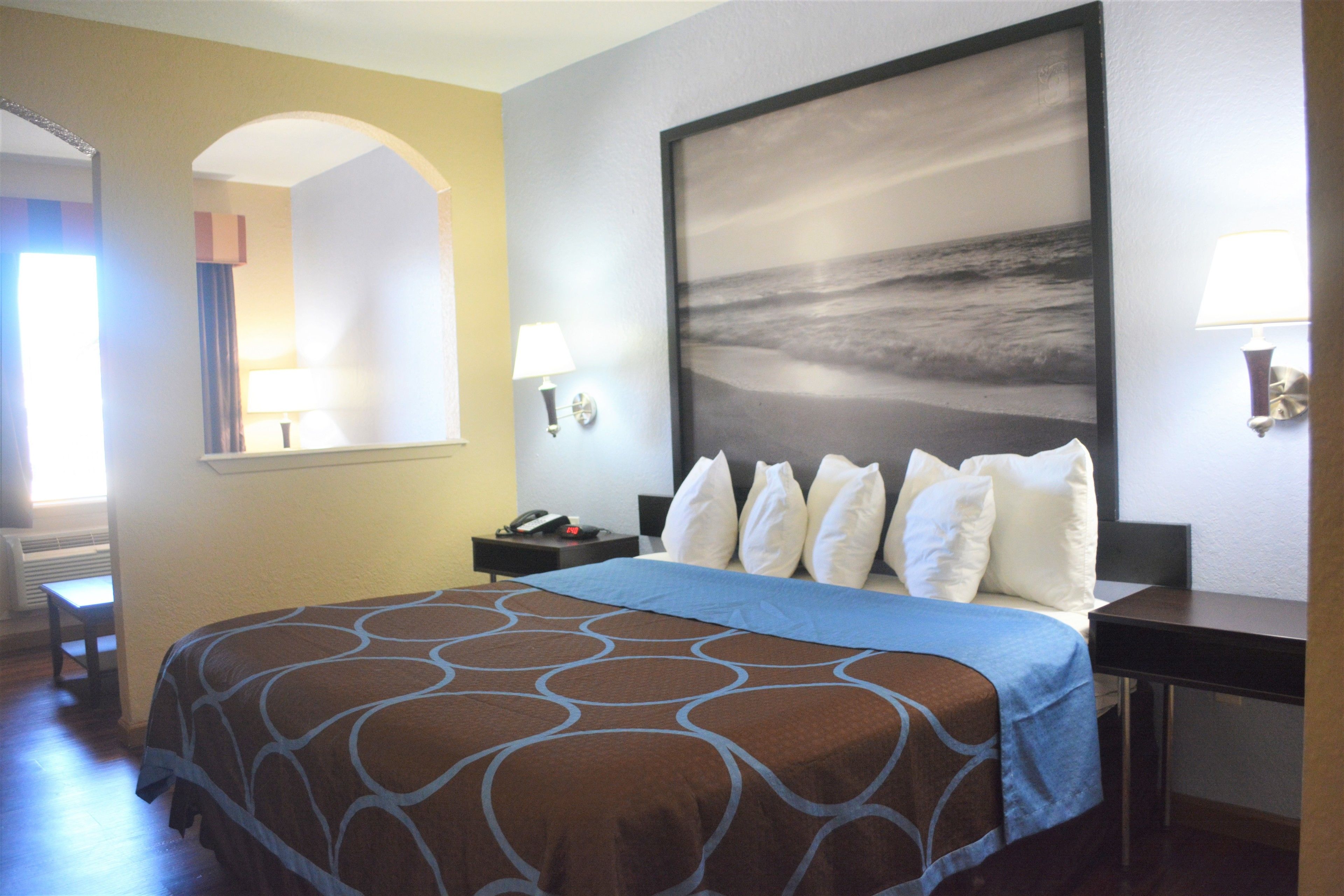 A hotel room with a large bed, a desk, a chair, a nightstand, a lamp, and a painting on the wall.