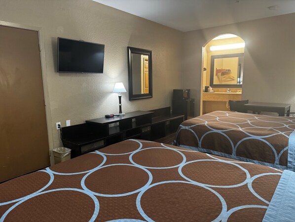 Habitación de hotel con dos camas, televisión, escritorio con lámpara, espejo y pequeña cocina.