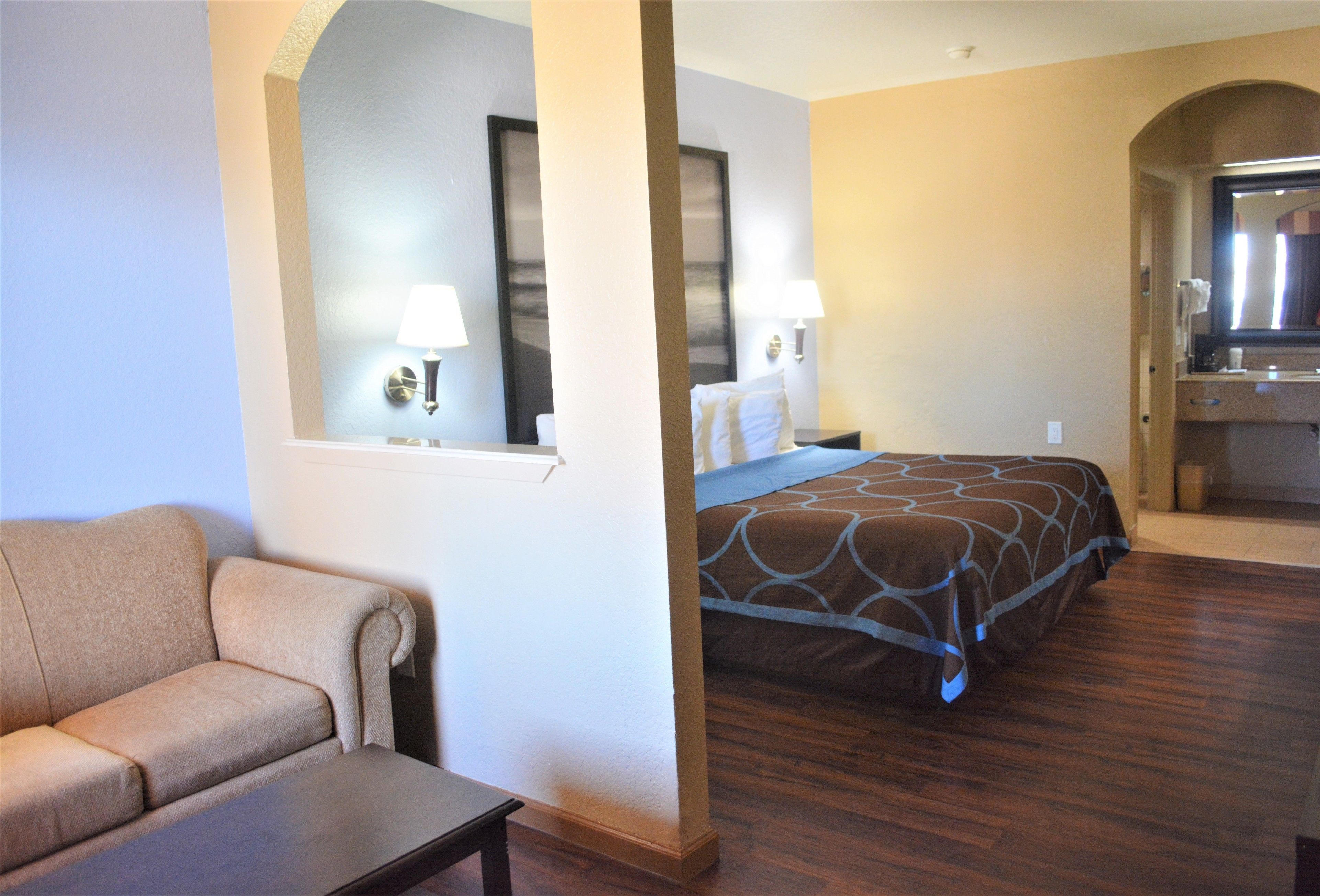 A hotel room with a sofa, a bed with a patterned bedspread, a nightstand with lamps, and a bathroom visible through an archway.