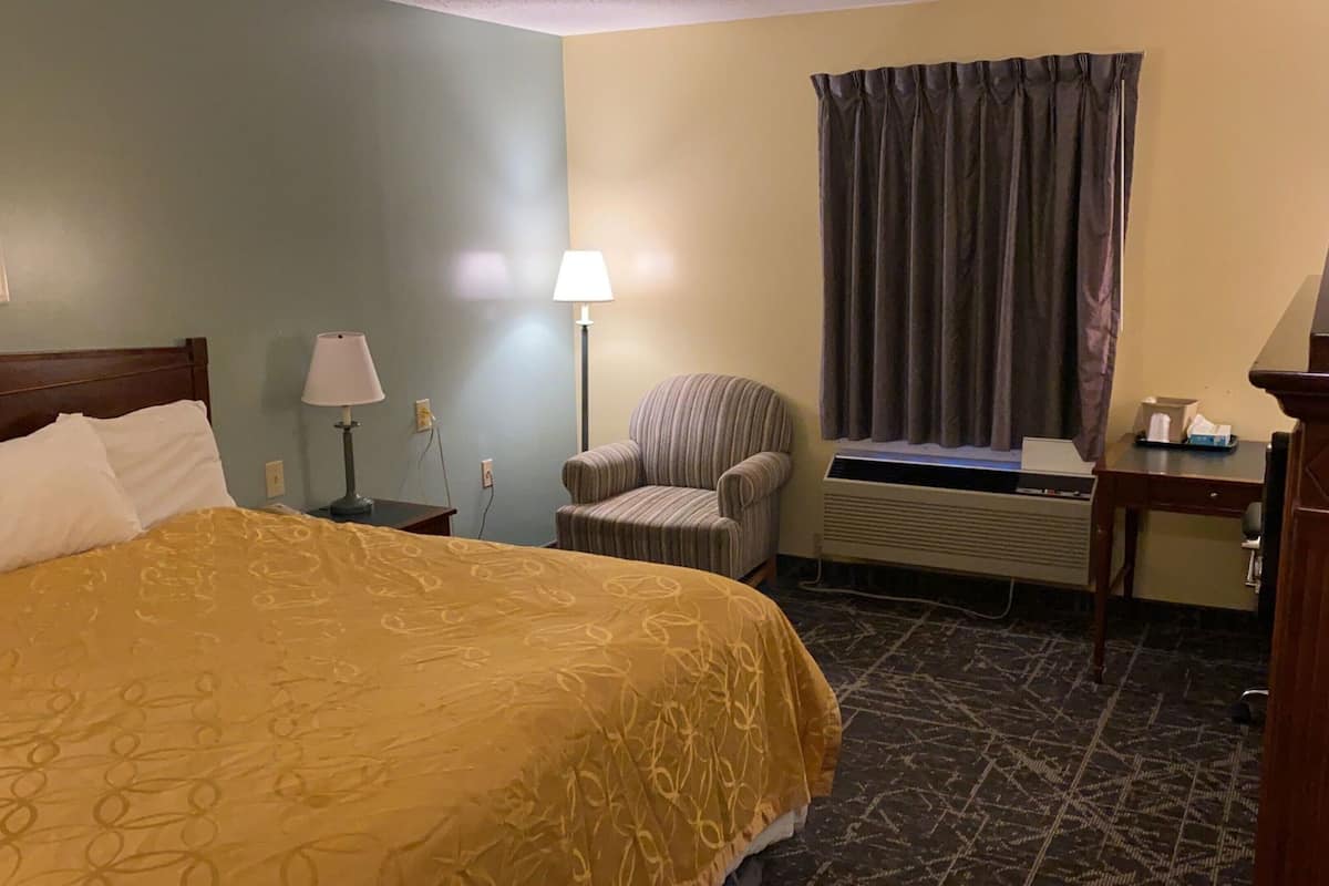 Quarto de hotel com cama, uma cadeira, escrivaninha, luminária e janela com cortinas.
