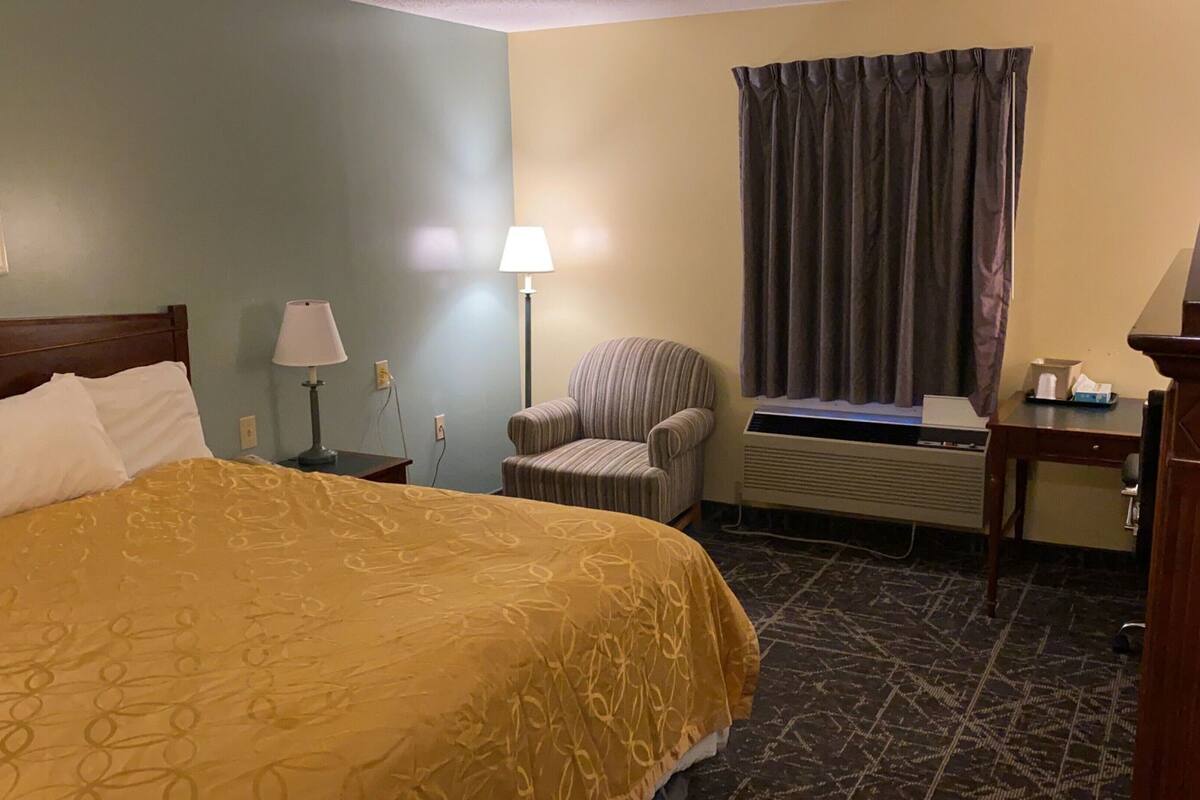 Quarto de hotel com cama, uma cadeira, escrivaninha, luminária e janela com cortinas.