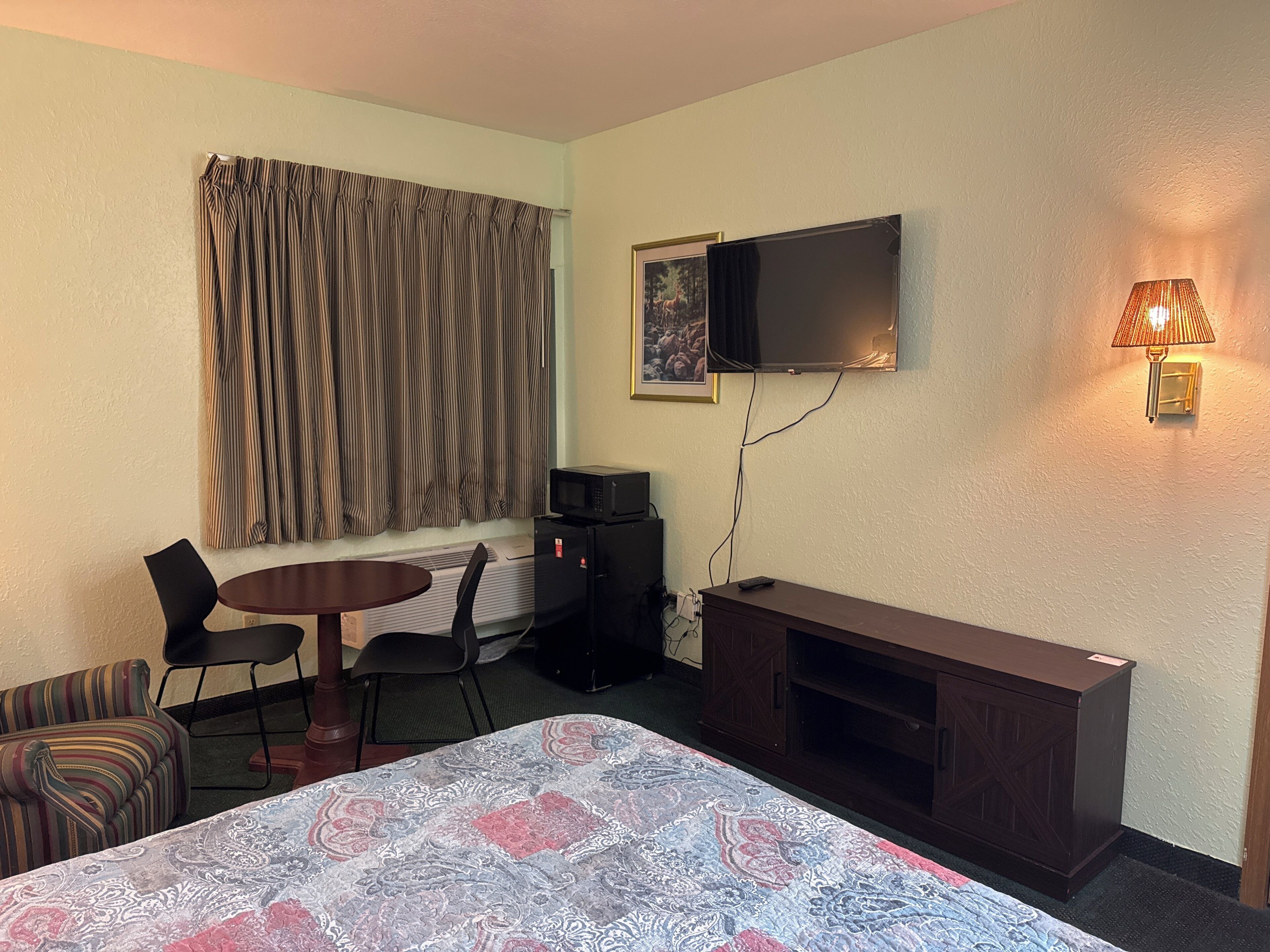 A hotel room with a bed, a desk with a chair, a television mounted on the wall, a microwave, and a lamp.
